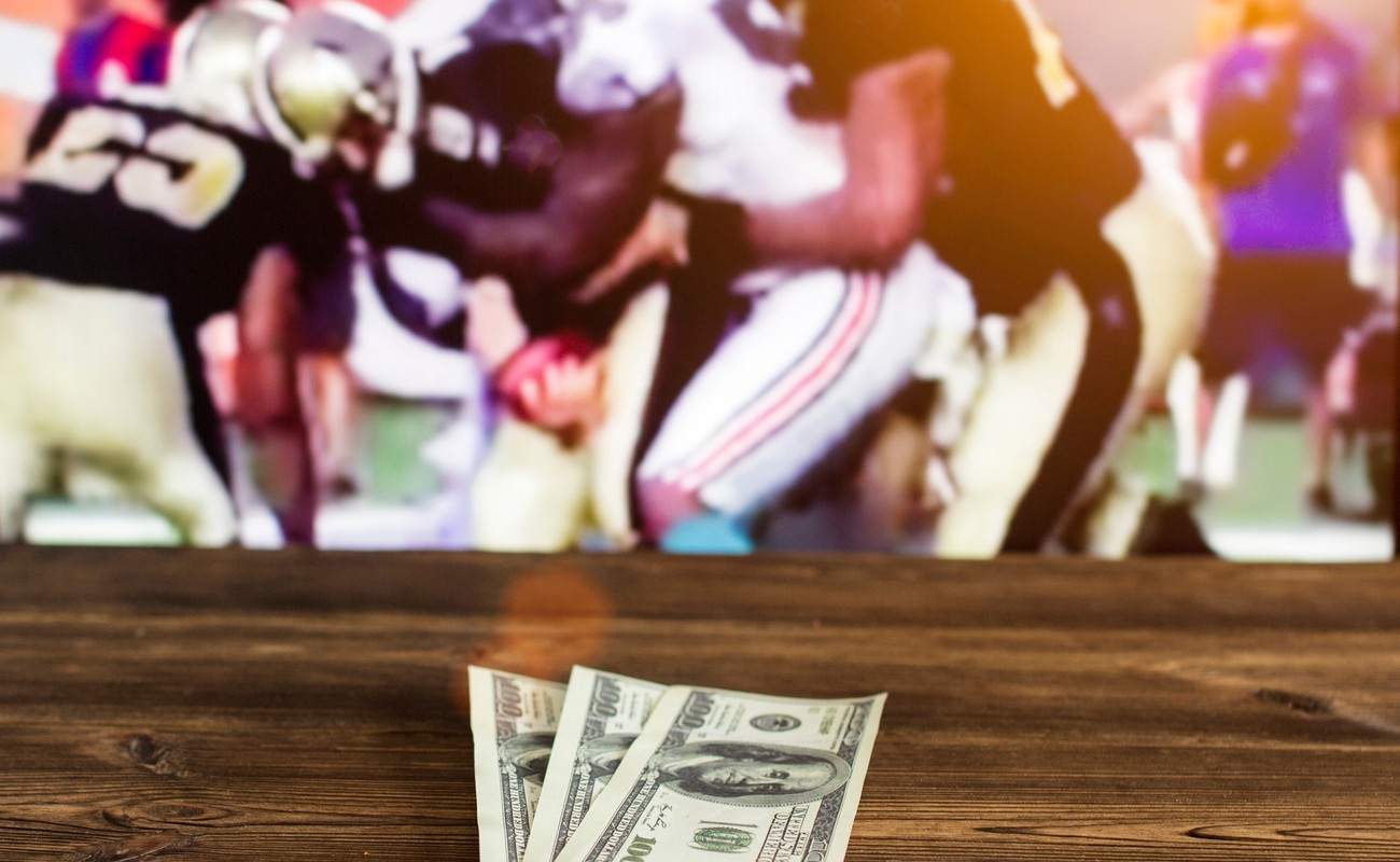 Dollars on a wooden table with a TV in the background showing American football