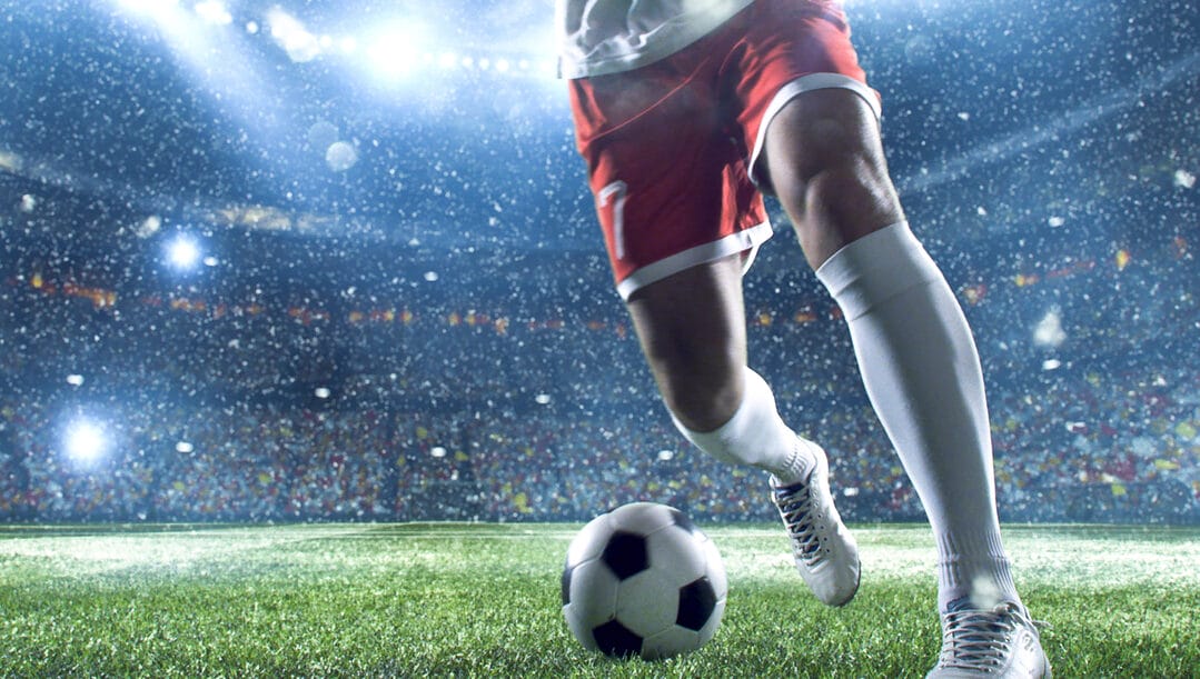 A close-up of a football player’s legs as he kicks the ball with the stadium stands behind him.
