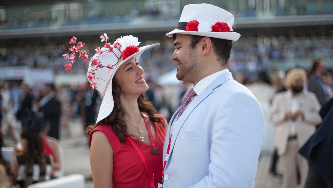 A well-dressed couple embrace and smile at each other at a horse racing event.