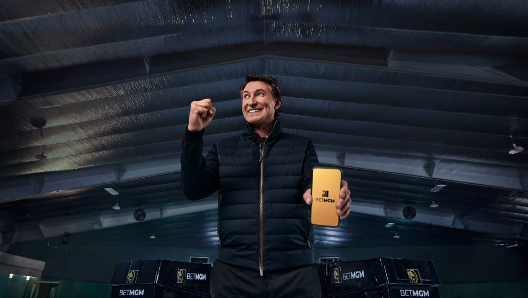 Wayne Gretzky in an ice hockey rink, holding a smart phone with the BetMGM app playing