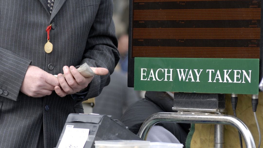 A racecourse bookmaker with cash in hand, next to a betting machine and a sign saying “Each Way Taken.”