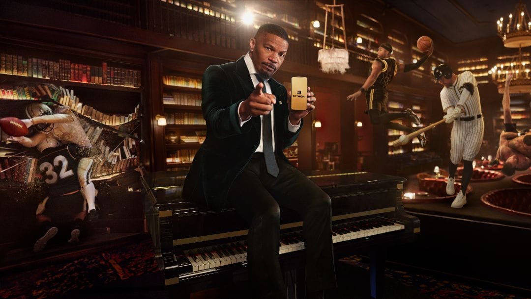 Jamie Foxx sitting on a piano while holding a mobile with the open BetMGM app